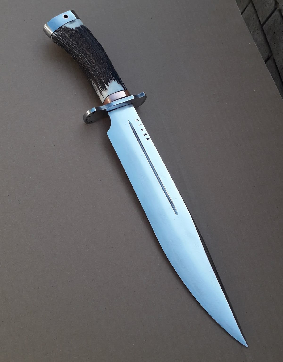 Handmade Bowie knife with dark stag horn handle and single fuller high carbon steel blade on gray surface