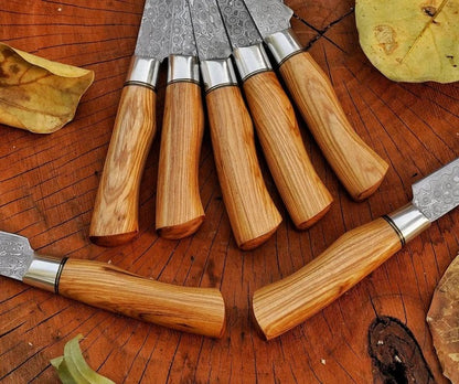 A set of kitchen knives with patterned steel blades and light brown wooden handles arranged on a rustic wooden surface with dried leaves.