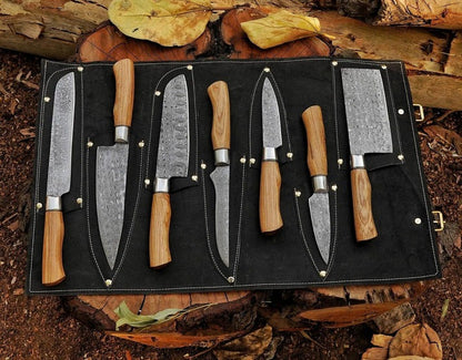 A 7-piece outdoor kitchen knife set with patterned steel blades and light wood handles, displayed in a black roll-up case on a log with autumn leaves.
