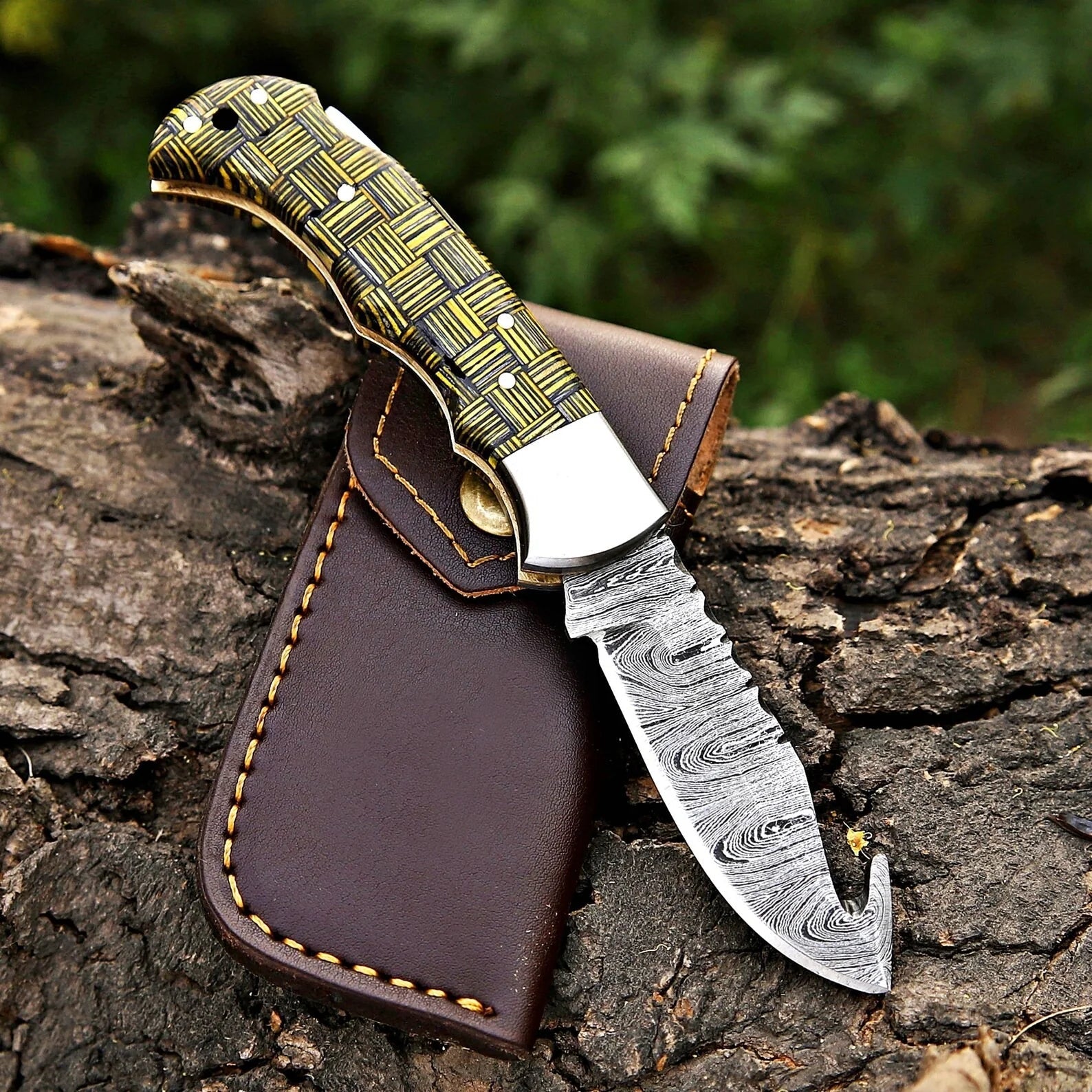 Handmade Damascus folding knife with woven composite handle and gut hook blade resting on dark brown leather sheath on rustic wood