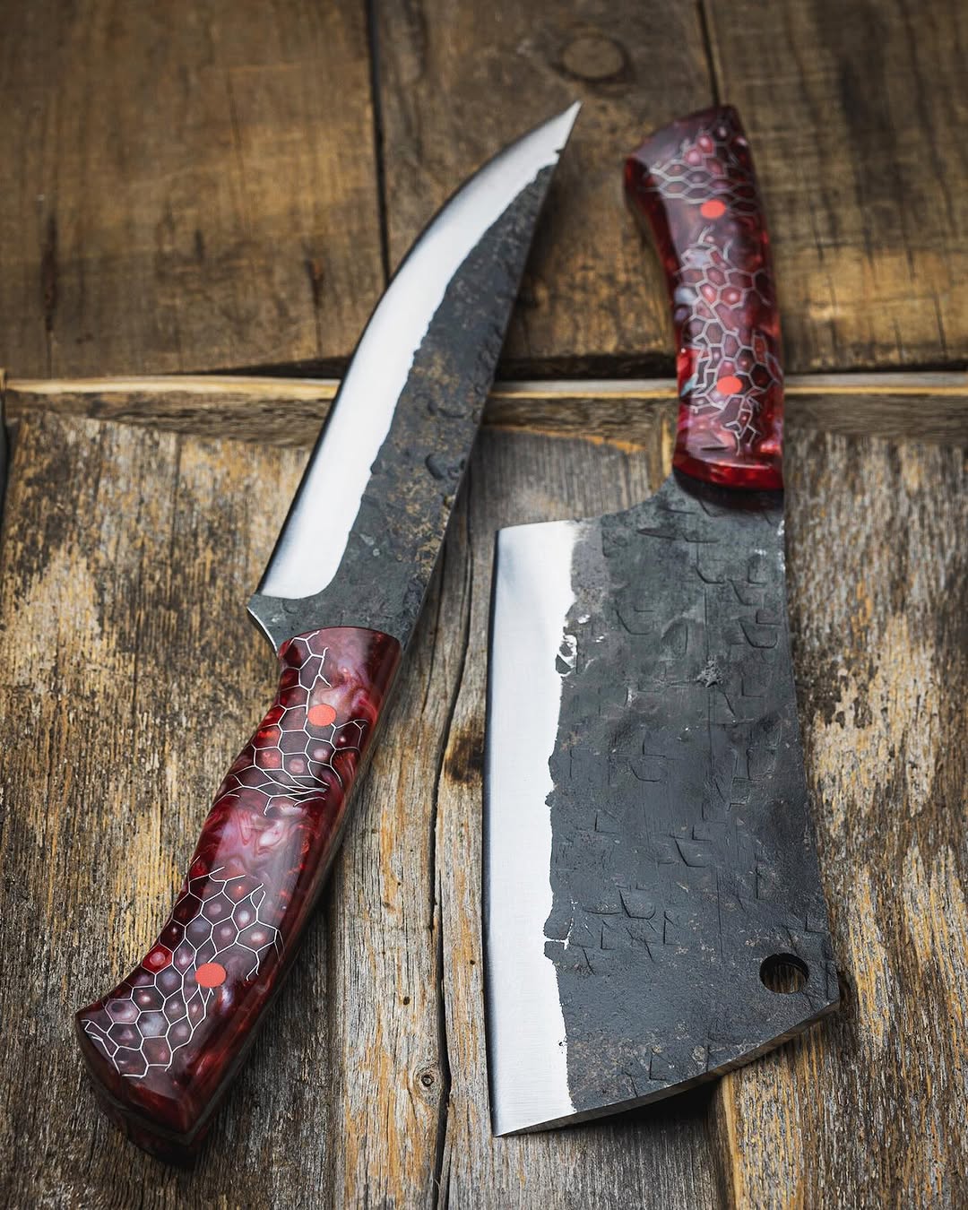 A handmade butcher knife and a cleaver with textured dark steel blades and red honeycomb-patterned handles, displayed on a rustic wooden background.