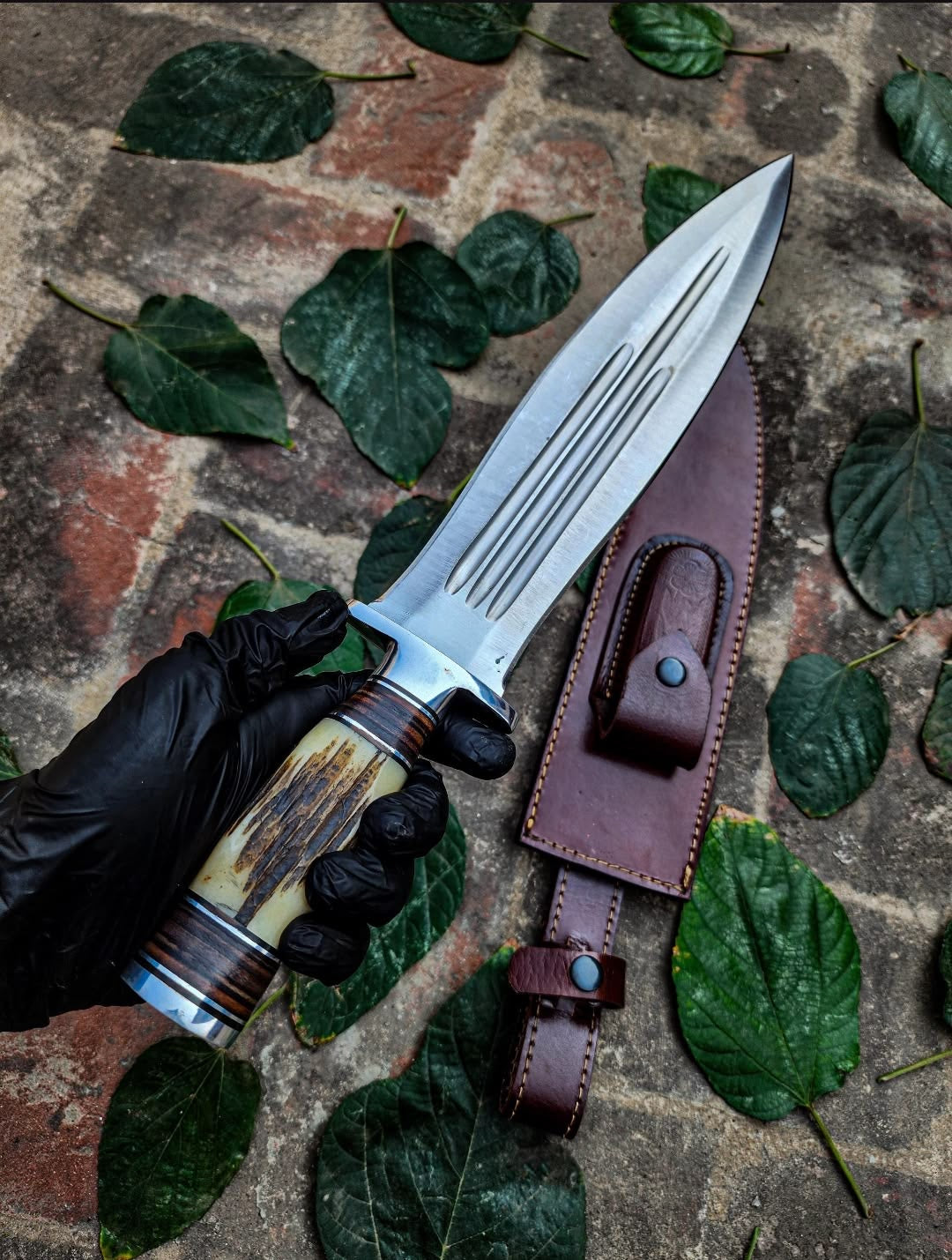 Handmade multi-fuller Bowie knife with stag horn and wood handle held in black glove with brown leather sheath on brick surface