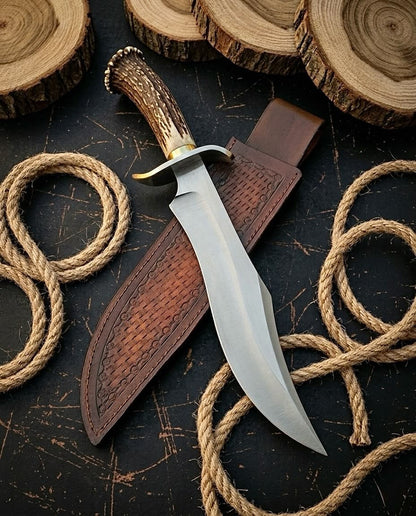 D2 steel Bowie knife with natural antler handle and brass guard beside embossed leather sheath on dark wood with rope