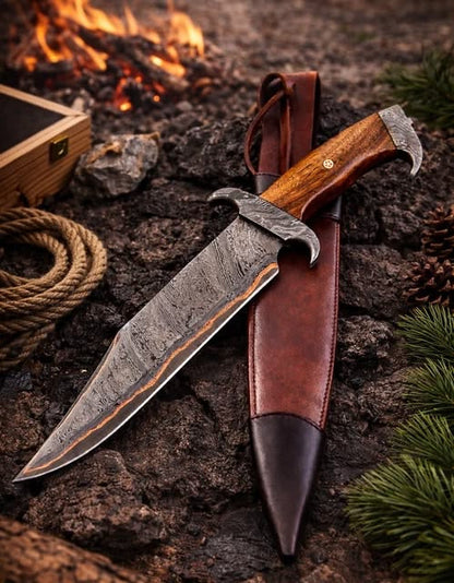 Handmade copper Damascus Bowie knife with walnut wood handle and eagle claw guard beside brown leather sheath on dark rocks with campfire background
