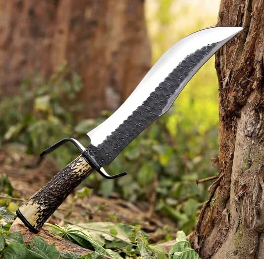 Handmade D2 steel Bowie knife with hammered spine antler handle and black D-guard stabbed into tree stump in forest