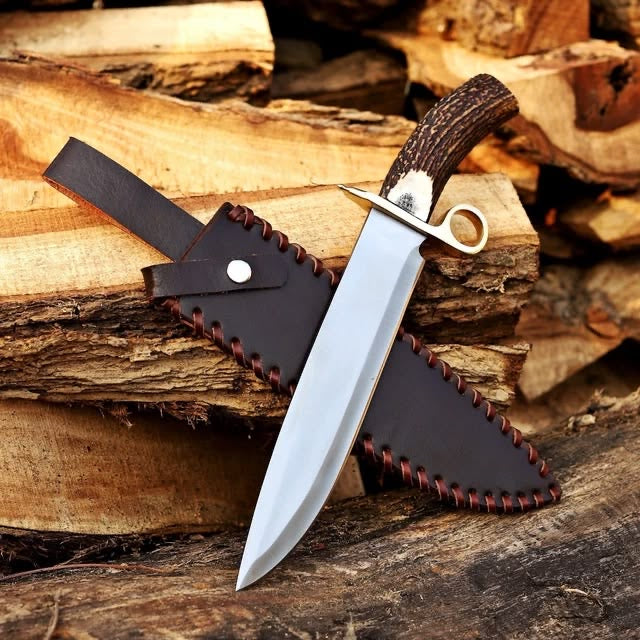Handmade D2 steel Bowie knife with mirror polished blade stag antler handle and brass guard resting on firewood with dark leather sheath