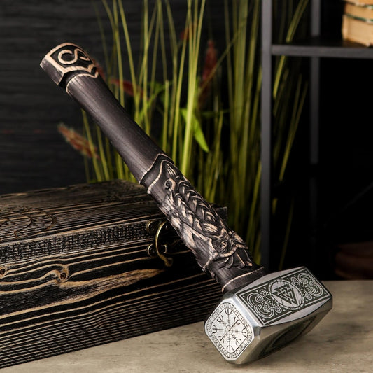 A decorative fantasy-style sword with an ornate dark metallic hilt and a silver-toned square blade featuring engraved symbols, displayed on a wooden surface with a dark background and tall artificial grass.