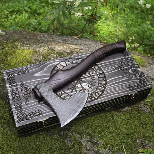 A dark wooden hatchet with a patterned steel blade rests on top of a matching carved wooden box outdoors on mossy ground with green plants in the background.