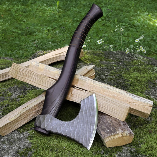 A tactical axe with a dark brown wooden handle and a textured silver steel head, resting on stacked wood logs outdoors on a mossy ground with green grass in the background.