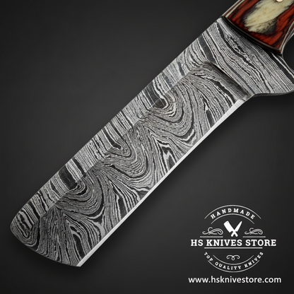 A close-up of a Damascus steel knife blade with a wavy gray and silver pattern, shown diagonally on a dark background. The handle is partially visible at the top right with a reddish-brown and cream inlay.