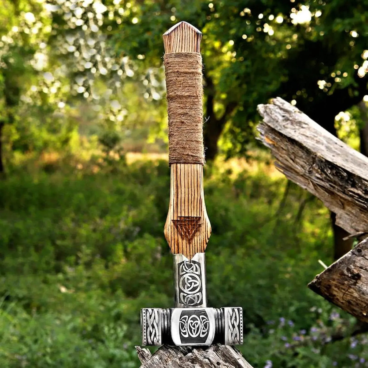 A Viking-style sword with a wooden handle wrapped in twine and a silver-colored metal hilt with Norse knotwork designs, standing upright on a log with a blurred forest background.