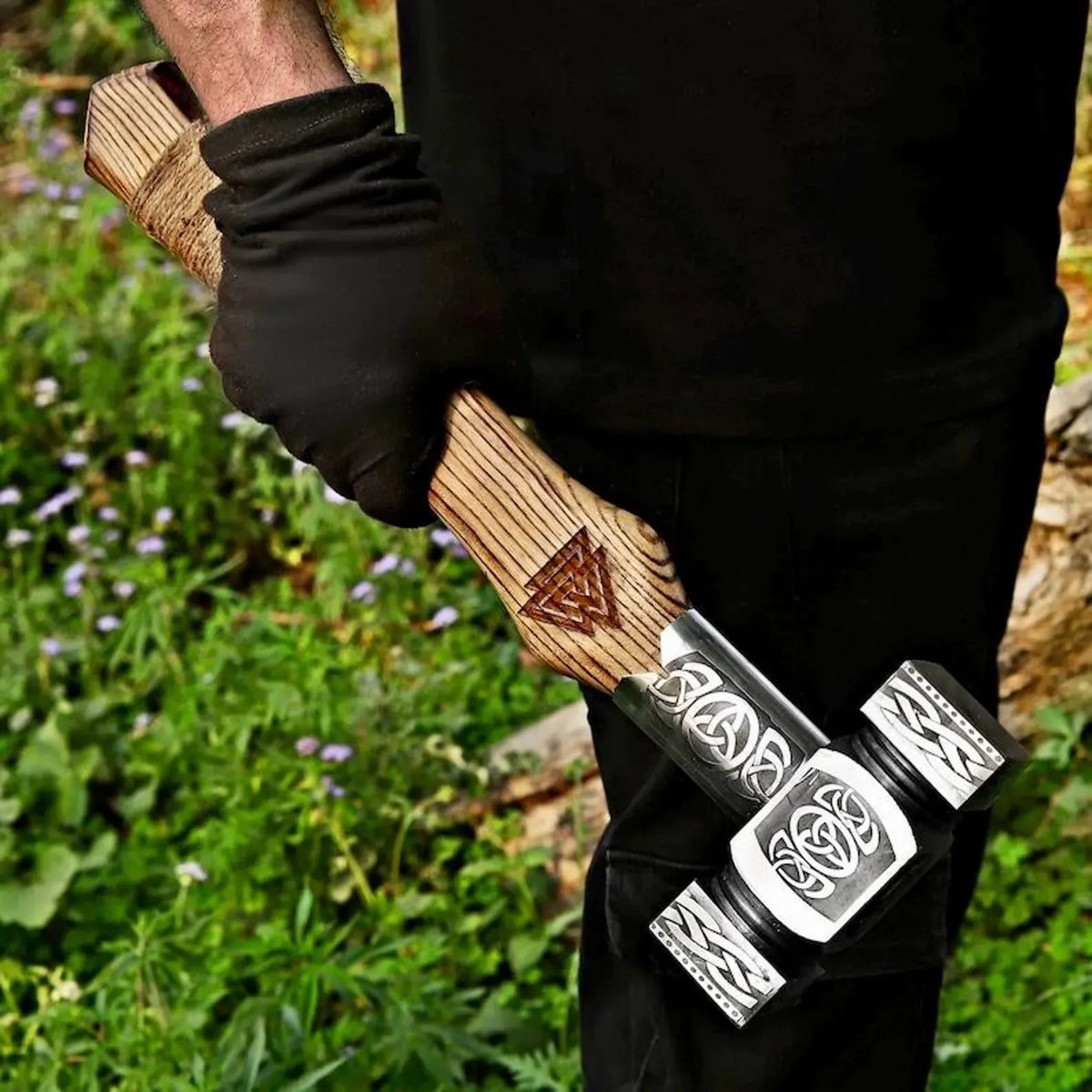A close-up of a person outdoors holding a hand axe/hatchet. The person is wearing a black glove and black clothing.
