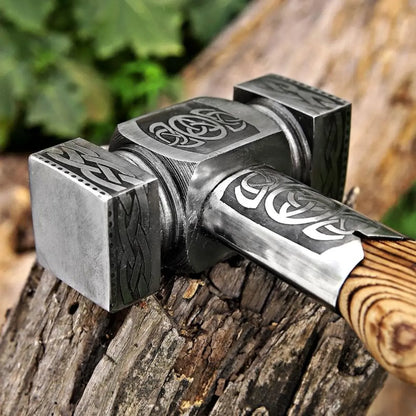 A close-up of a hand-forged Viking-style hammer resting on a rustic log, featuring an engraved steel head and a light brown wooden handle.