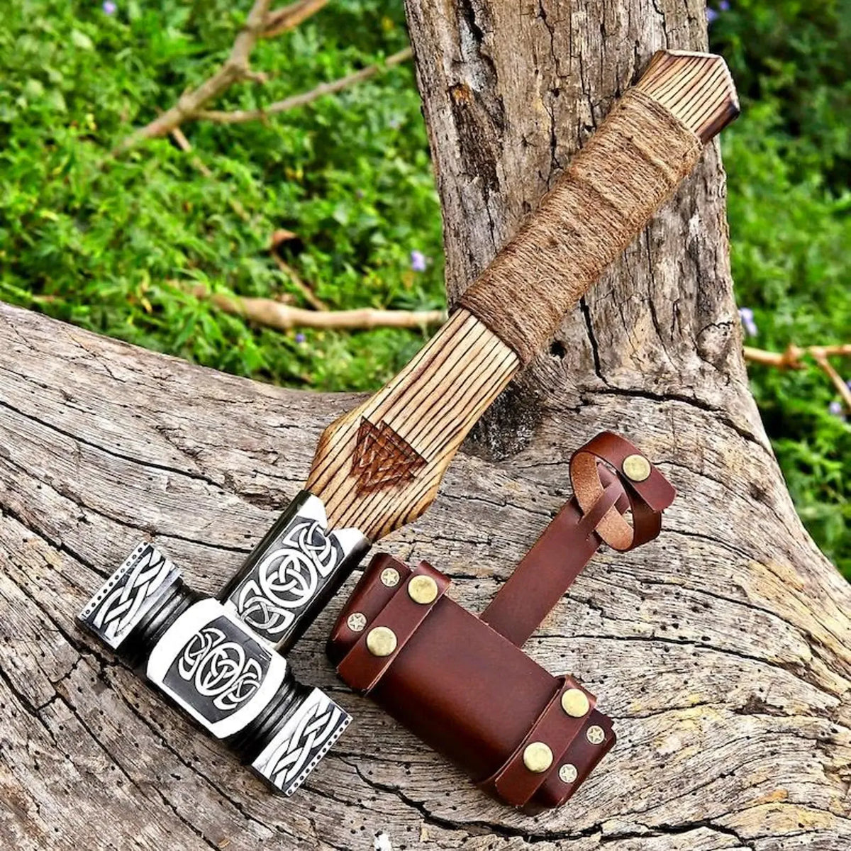 A handcrafted Viking-style bearded axe with a striped wooden handle, black and silver engraved metal head, and a brown leather sheath, displayed on a weathered tree trunk with green foliage in the background.