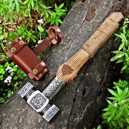 A handcrafted Viking-style bearded axe with a carved wooden handle and engraved steel head, displayed on a weathered log with a brown leather sheath and green foliage in the background.