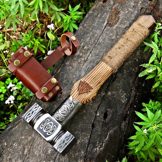 A handcrafted Viking-style bearded axe with a carved wooden handle and engraved steel head, displayed on a weathered log with a brown leather sheath and green foliage in the background.