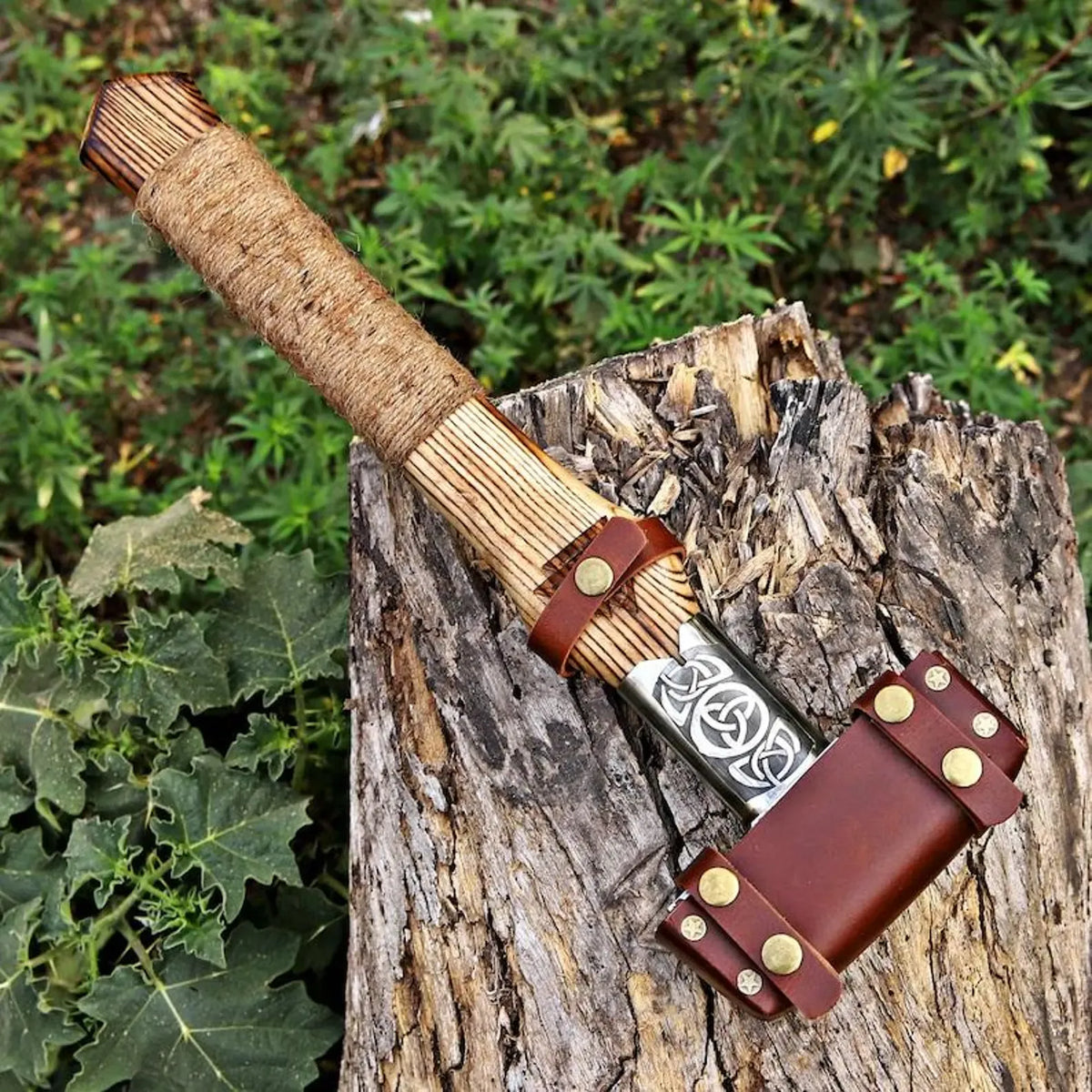 A handcrafted Viking-style axe with a natural wood handle, silver patterned steel blade, and brown leather sheath, resting on a weathered tree stump with green foliage in the background.