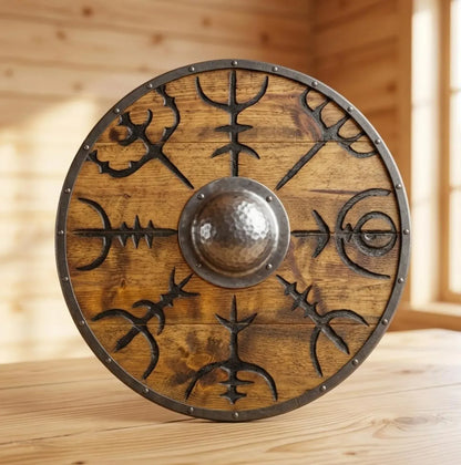 A round wooden Viking-style shield with engraved black runic symbols and a hammered metal center, displayed on a wooden surface against a wood-paneled background.