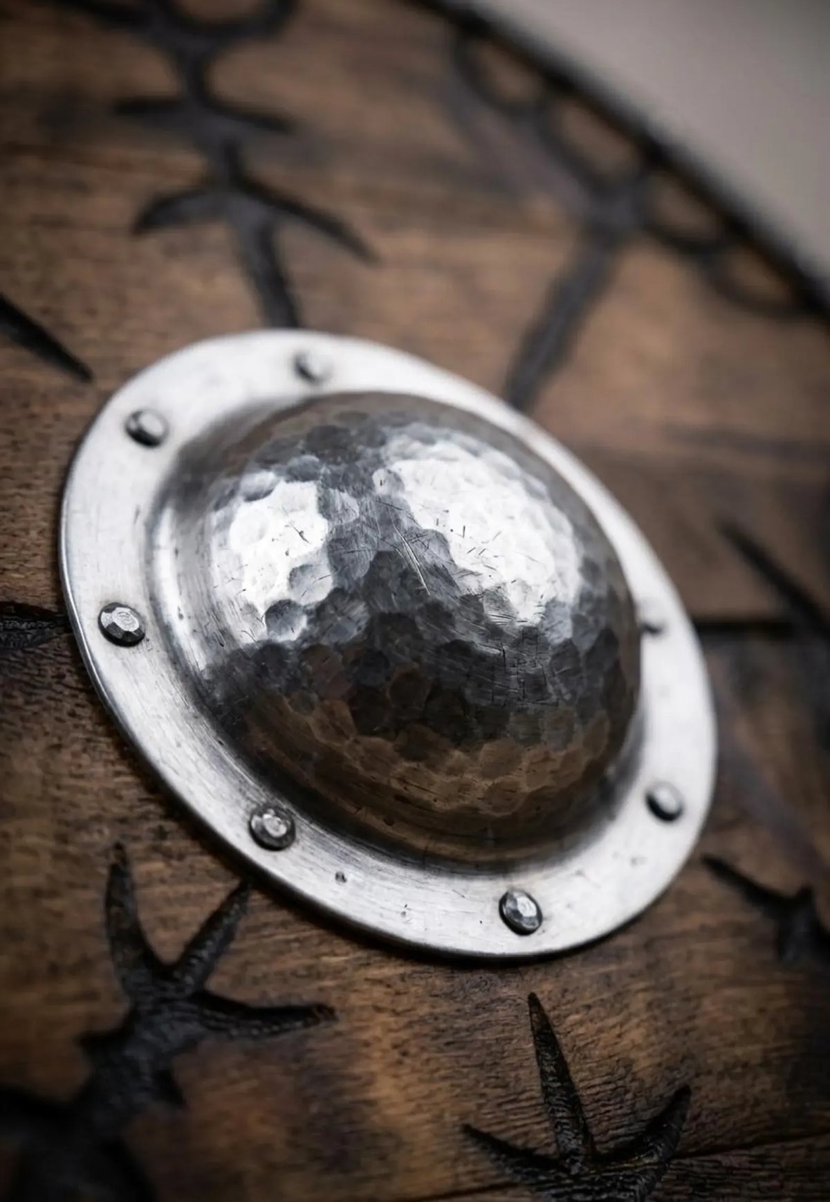 Close-up of a hammered metal round shield boss mounted on a dark wooden shield with black engraved star-like patterns.