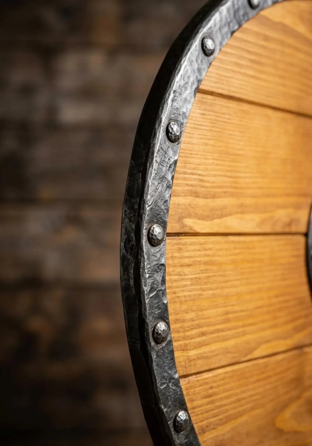 Close-up of a wooden shield or wall decor piece with a warm brown wood grain surface and a dark metal rim secured by round rivets, set against a blurred dark rustic background.