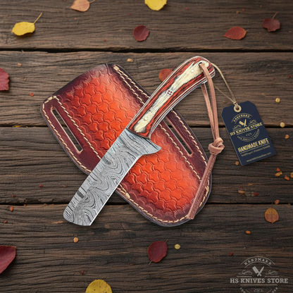 A Damascus steel fixed-blade knife with a textured orange-brown leather sheath, displayed on a dark wooden surface with scattered autumn leaves. A blue brand tag for 'HS Knives Store' is attached to the sheath.