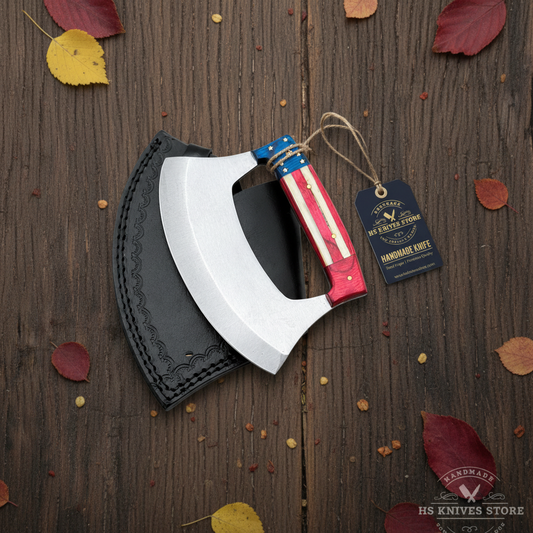 D2 steel ulu knife flat lay with American flag handle hang tag and black embossed leather sheath on dark rustic wood with autumn leaves
