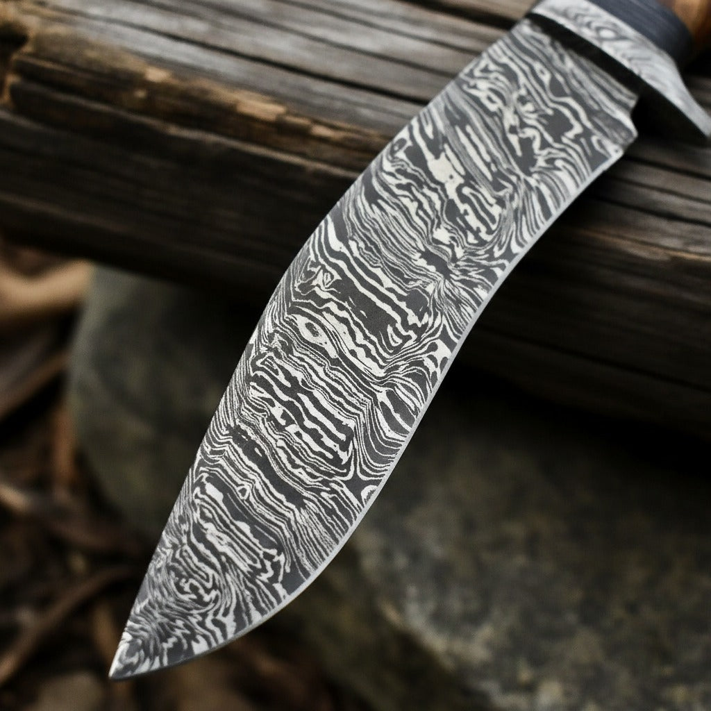 A close-up of a Damascus steel knife blade with a distinctive wavy pattern, resting against a rustic dark wooden log outdoors.