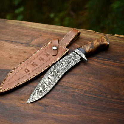 A fixed-blade knife with a patterned steel blade and a brown wood-grain handle, placed on a dark wooden surface next to a brown leather sheath.