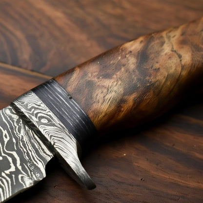 Close-up of a knife with a patterned Damascus-style blade and a richly figured brown wood handle, resting on a dark wood surface.
