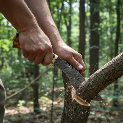 A close-up action shot of a person's hands using a large machete to cut a tree branch in a forest. The machete has a textured gray blade with a black and white wavy pattern, a light-colored handle, and a yellow accent near the guard.