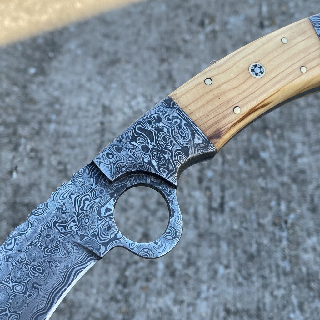 knife with a circular hole in the blade, resting on a textured gray concrete surface. The knife features a patterned steel blade, a dark steel bolster, and a light-colored wood handle.