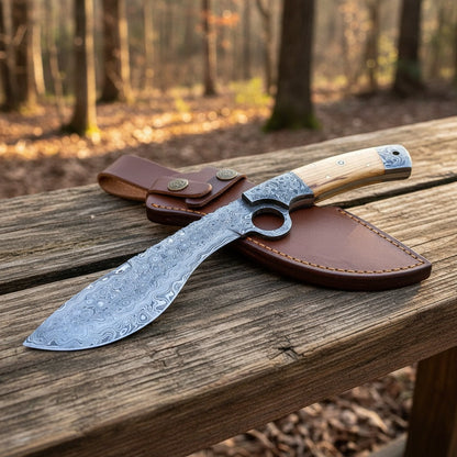 A fixed-blade knife with a patterned steel blade, a light wood handle, and a silver metal guard, displayed on a brown leather sheath on a rustic wooden bench with a blurred forest background.