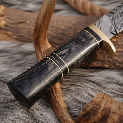  close-up of a knife with a dark wood handle, patterned steel blade, and gold-toned guard, resting on a fur surface with antlers and wood logs as props.