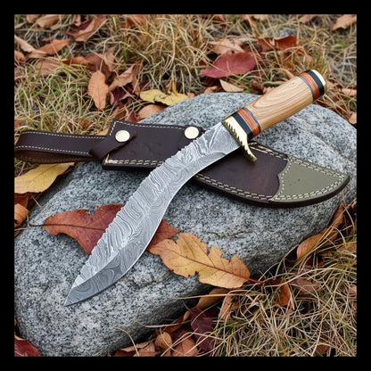fixed-blade knife with a patterned steel blade, a light wood handle, and a brass guard, displayed on a dark brown leather sheath over a gray rock outdoors on grass with fallen leaves.