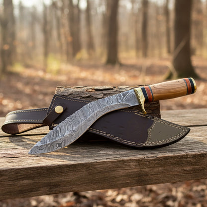  Damascus steel fixed-blade knife with a patterned steel blade, a light wood handle with colorful stripes, and a brass bolster, resting on a dark brown leather sheath over a log in an outdoor forest setting.