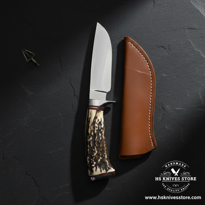 D2 steel skinner hunting knife displayed vertically on dark slate with stag handle and tan leather sheath