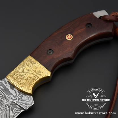 Macro view of dark brown wood handle with black pins ruby pin and engraved brass bolster and leather lanyard on Damascus tracker knife