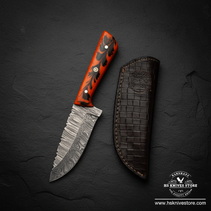 Damascus steel skinner knife displayed vertically on dark slate with orange resin handle and crocodile leather sheath