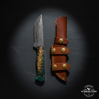 Damascus steel skinner knife displayed vertically on dark slate with green resin pine cone handle and tan leather belt sheath luxury shot