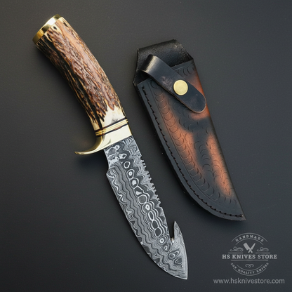 Damascus steel gut hook hunting knife stag antler handle brass collar bolster dark black fish scale leather sheath dark charcoal studio background