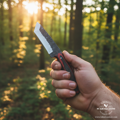 One hand gripping carbon steel drop point skinner knife with red black pakkawood handle in outdoor forest golden hour lifestyle setting