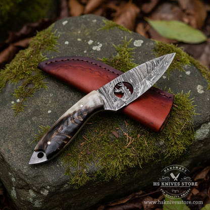 Damascus steel skinner knife ram horn handle resting on mossy rocks in outdoor forest setting with dark burgundy leather sheath