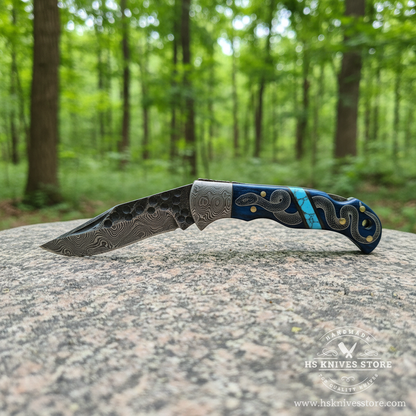 Damascus steel folding pocket knife open blade resting on granite rock in bright outdoor forest setting clear Damascus pattern visible