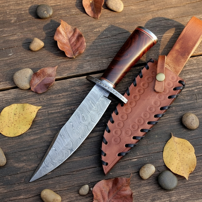 Handmade Damascus Bowie knife flat lay on rustic wood with brown leather sheath and black lace stitching