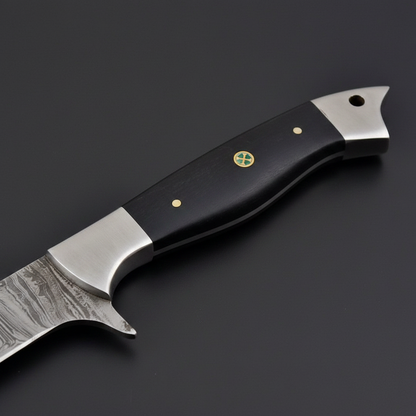 Close-up macro of black micarta handle with brass pins, green cross medallion, steel bolster finger notch and pommel cap