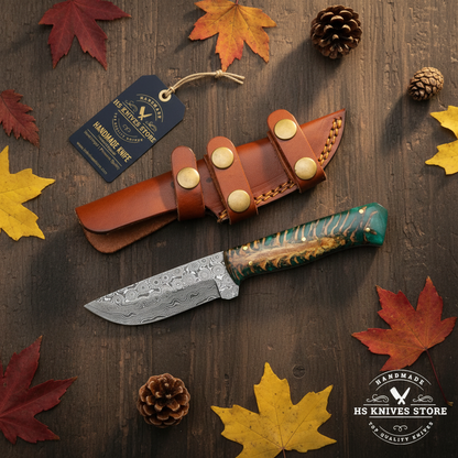Damascus skinner knife flat lay with green resin pine cone handle and HS Knives Store hang tag on rustic dark wood with autumn leaves