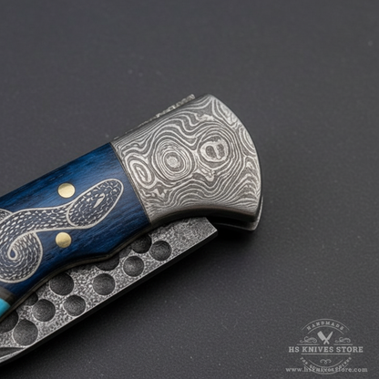 Close-up macro of Damascus steel bolster with hammered dimple texture and blue wood snake engraved handle turquoise inlay on folding knife