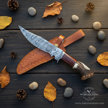 Damascus steel Bowie knife stag crown handle flat lay rustic dark wood surface with autumn leaves and pine cones