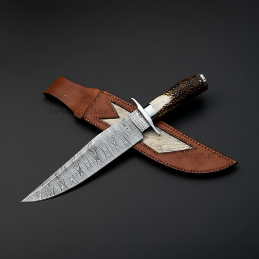 Hand forged Damascus Bowie knife with natural stag horn handle and steel cross guard on dark charcoal gray studio background right facing