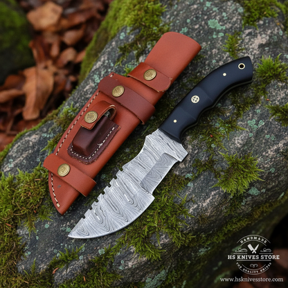 Damascus steel tracker knife black micarta handle resting on mossy rocks in outdoor forest setting with cognac leather sheath